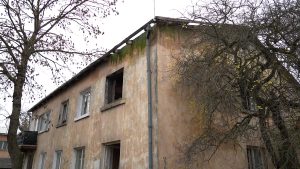 Pamests divstāvu nams ar dzeltenīgu apmetumu, bojātu jumtu un izsistiem logiem.