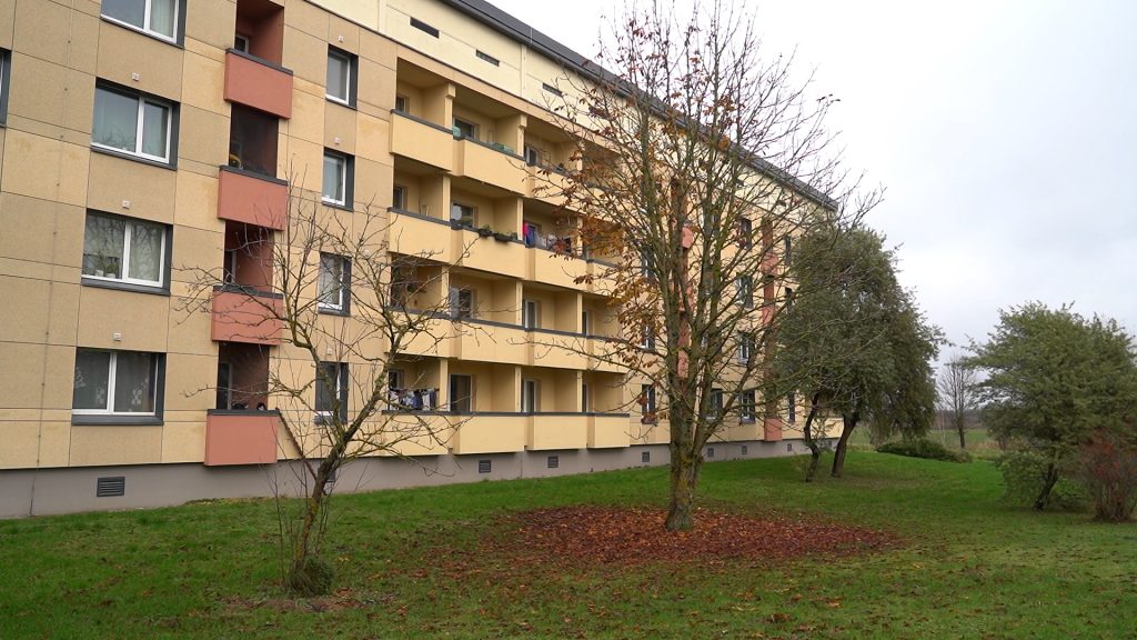 Dzīvokļu ēka dzeltenīgi oranžā tonī ar balkoniem un kokiem priekšplānā.