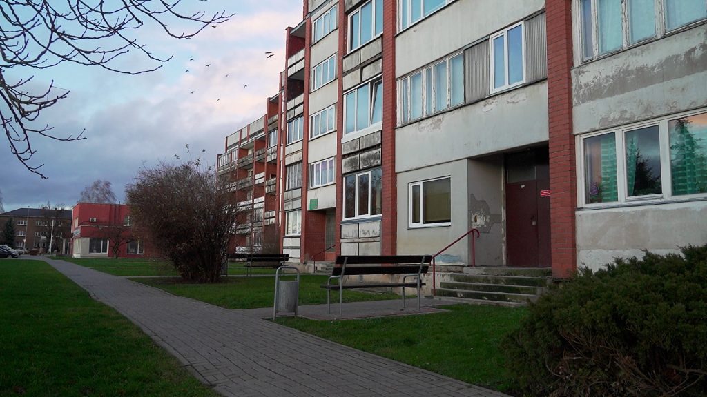Padomju laika daudzdzīvokļu nams ar soliņiem un celiņu priekšplānā.