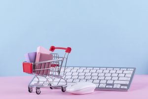 Miniatūrs iepirkumu ratiņš ar dāvanu kastītēm pie datora klaviatūras un peles, simbolizējot iepirkšanos internetā.