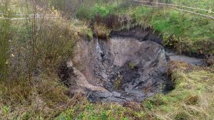 Dziļš nogruvums ar izskalotu augsni un akmeņiem, norobežots ar drošības lentēm.