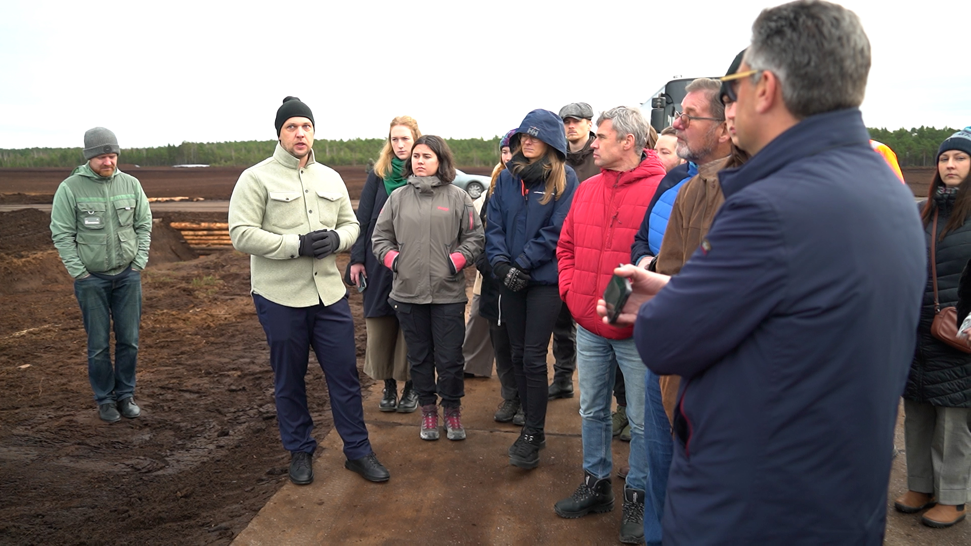 Cilvēku grupa (uzņēmēji, zinātnieki) klausās runātājā, stāvot uz dēļu laipas Drabiņu kūdras purvā.