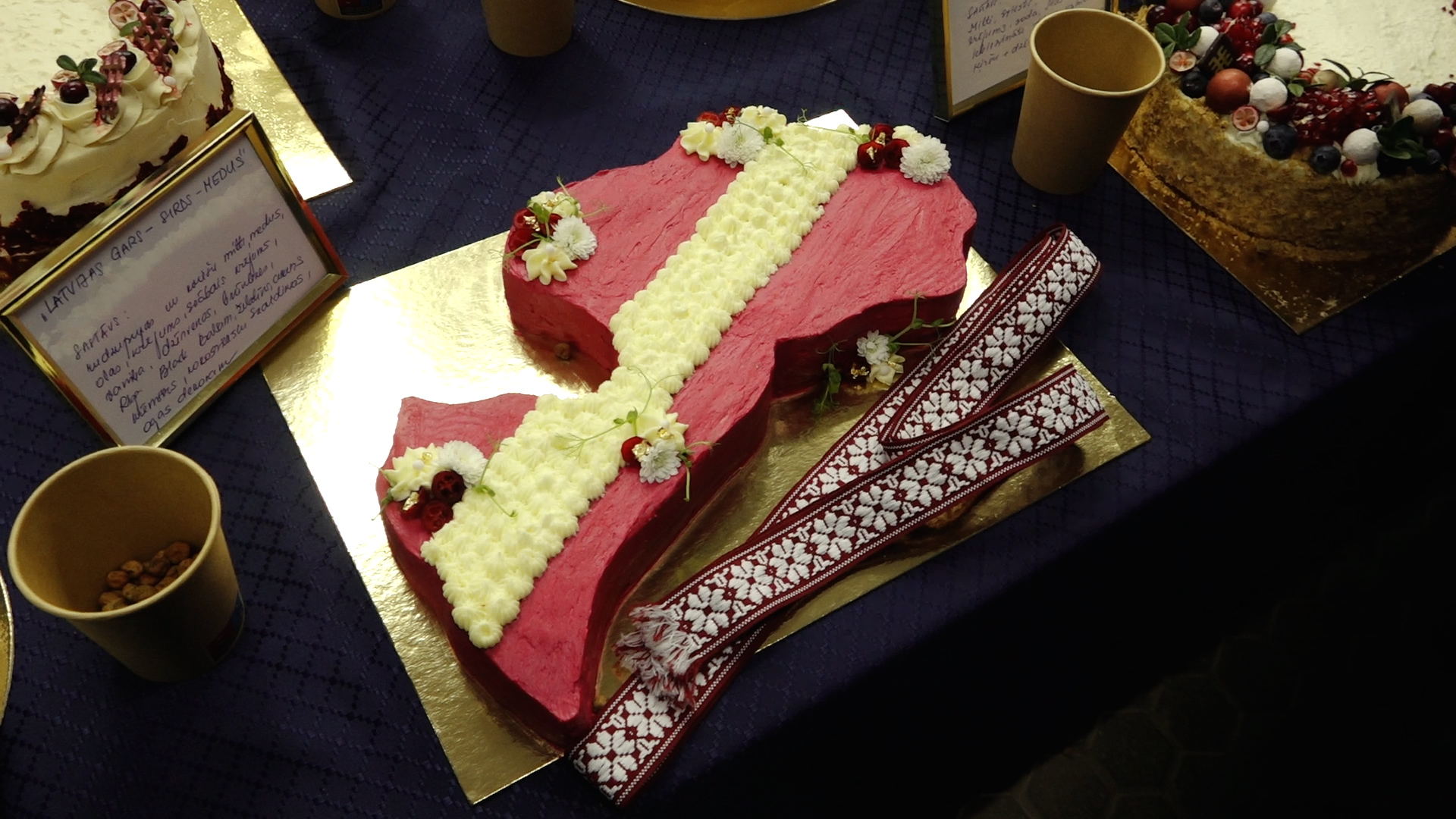 Torte Latvijas kontūras formā ar sarkanbaltsarkanu dekoru un tautisku jostu.