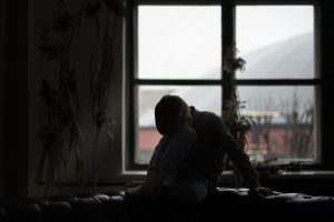 Bērns sēž uz dīvāna pie loga, galvu noliecis pret ceļiem, tumšā un klusā telpā.