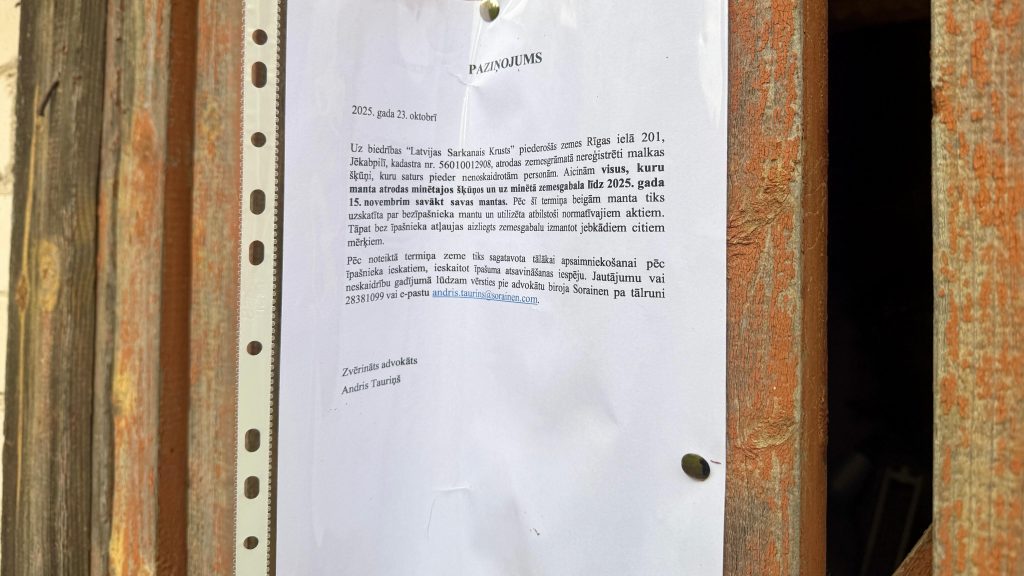 Tuvplāna fotogrāfija ar advokāta Andri Tauriņa paziņojumu, kas izdrukāts uz A4 lapas, par Latviju Sarkanajam Krustam piederošu zemi Jēkabpilī un aicinājumu savākt mantas no šķūņiem līdz 2025. gada 15. novembrim.