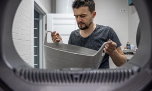 Vīrietis ar veļas grozu, skatoties caur veļas mazgājamās mašīnas atveri.