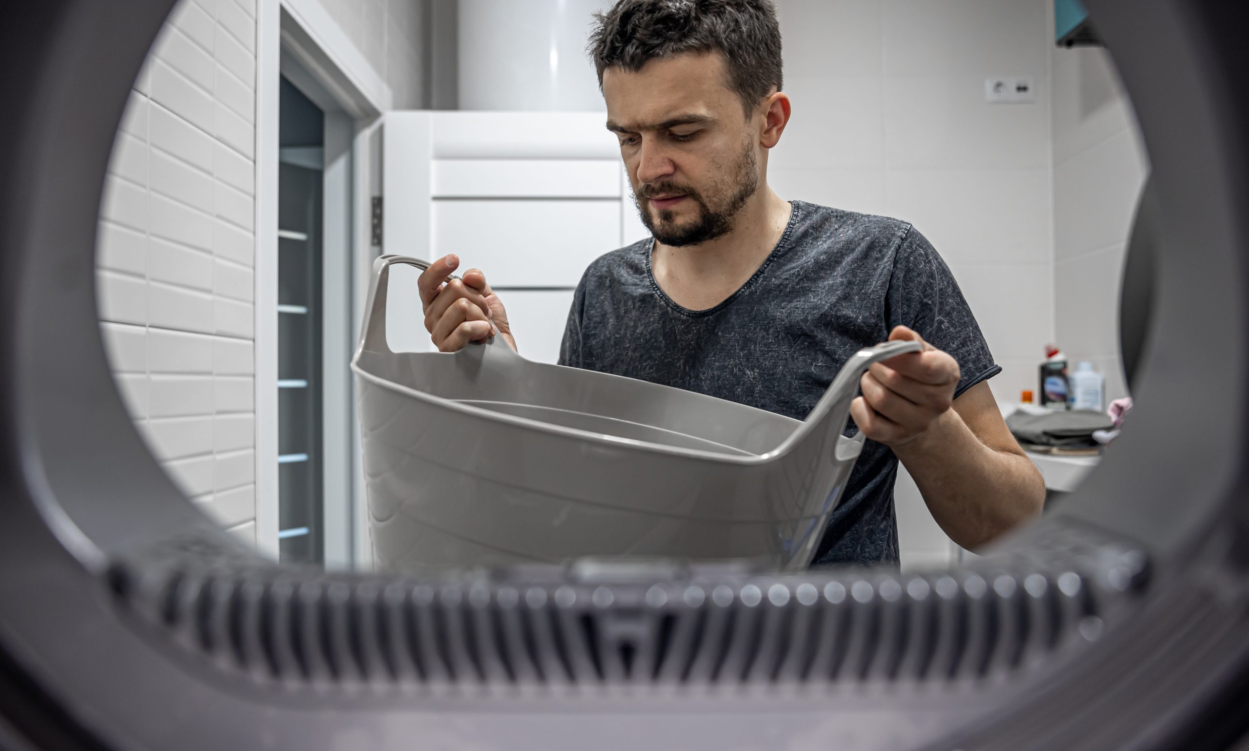 Vīrietis ar veļas grozu, skatoties caur veļas mazgājamās mašīnas atveri.
