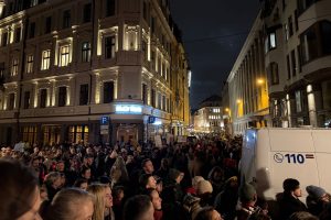 Liels cilvēku pūlis tumšā ielā Rīgas centrā, protestējot pret izstāšanos no Stambulas konvencijas. Ielas malā redzams policijas mikroautobuss.