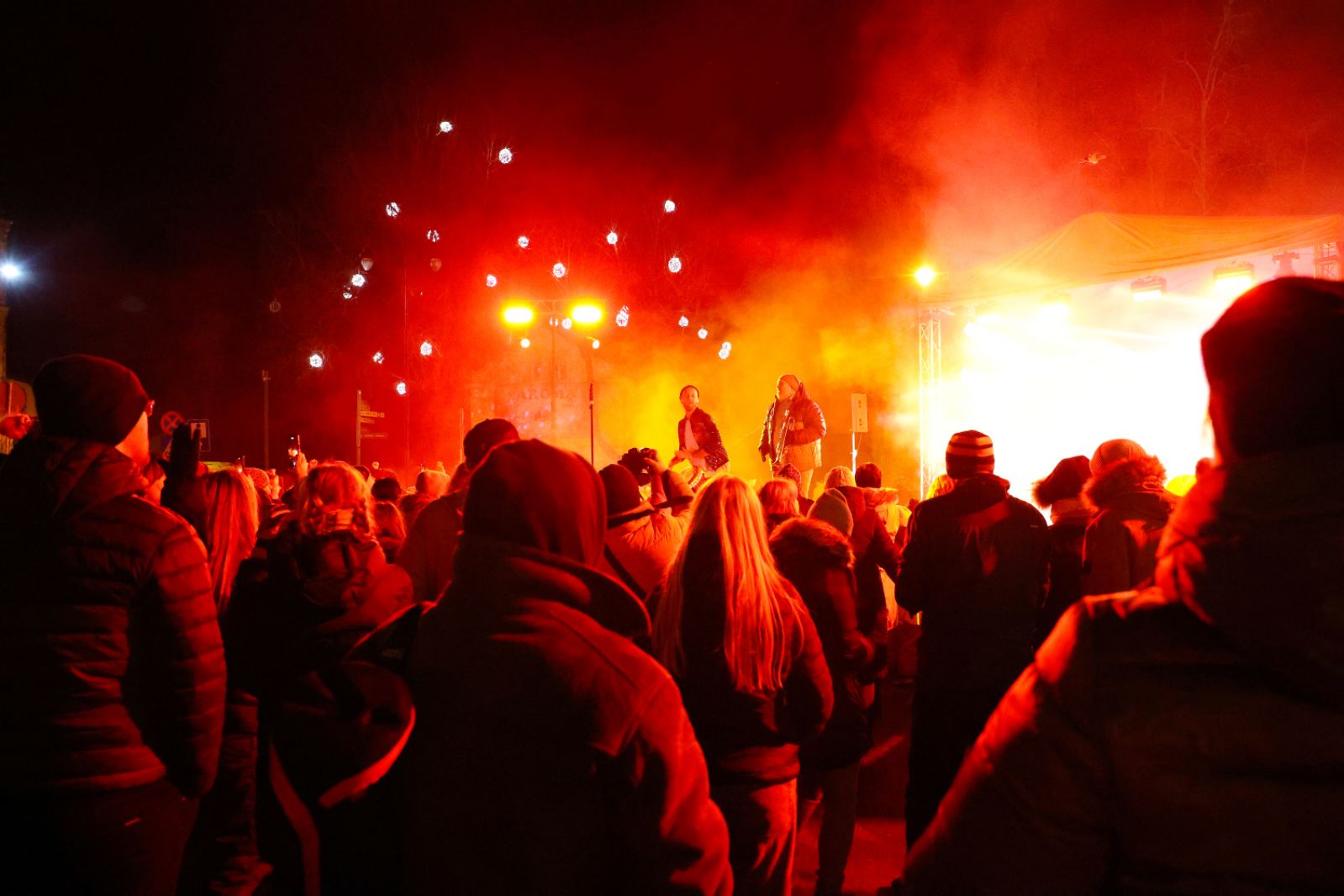 Cilvēku pūlis nakts pasākumā Cēsīs, vērojot skatuvi, ko apgaismo spilgti sarkani un dzelteni gaismas efekti.