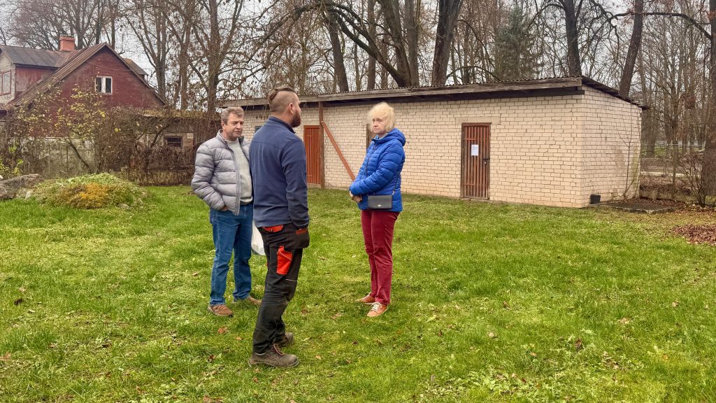 Trīs cilvēki (divi vīrieši un viena sieviete, iespējams, LSK pārstāve Velta Ozoliņa un iedzīvotāji) stāv uz zaļa zāliena pie ķieģeļu šķūņiem un sarunājas.