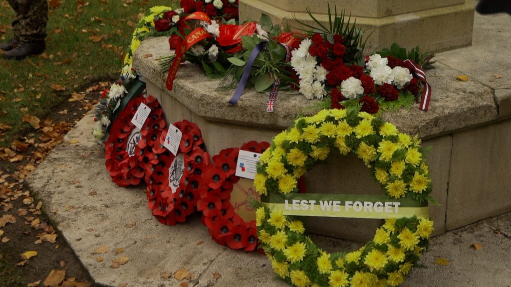 Tuvplāns pie pieminekļa noliktiem vainagiem, tostarp sarkano magoņu un dzelteno ziedu vainags ar uzrakstu "Lest We Forget"