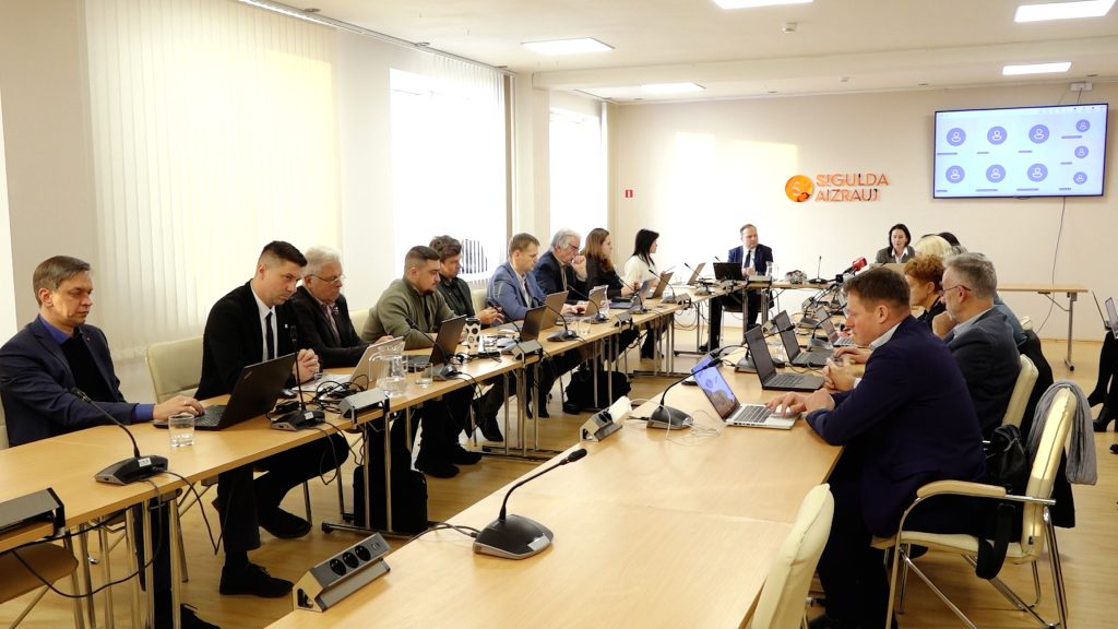 Plašs skats uz Siguldas novada domes sēdi, deputāti sēž pie galdiem ar klēpjdatoriem.