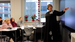 Skolotāja vada stundu modernā klasē, skolēni klausās un veic pierakstus.