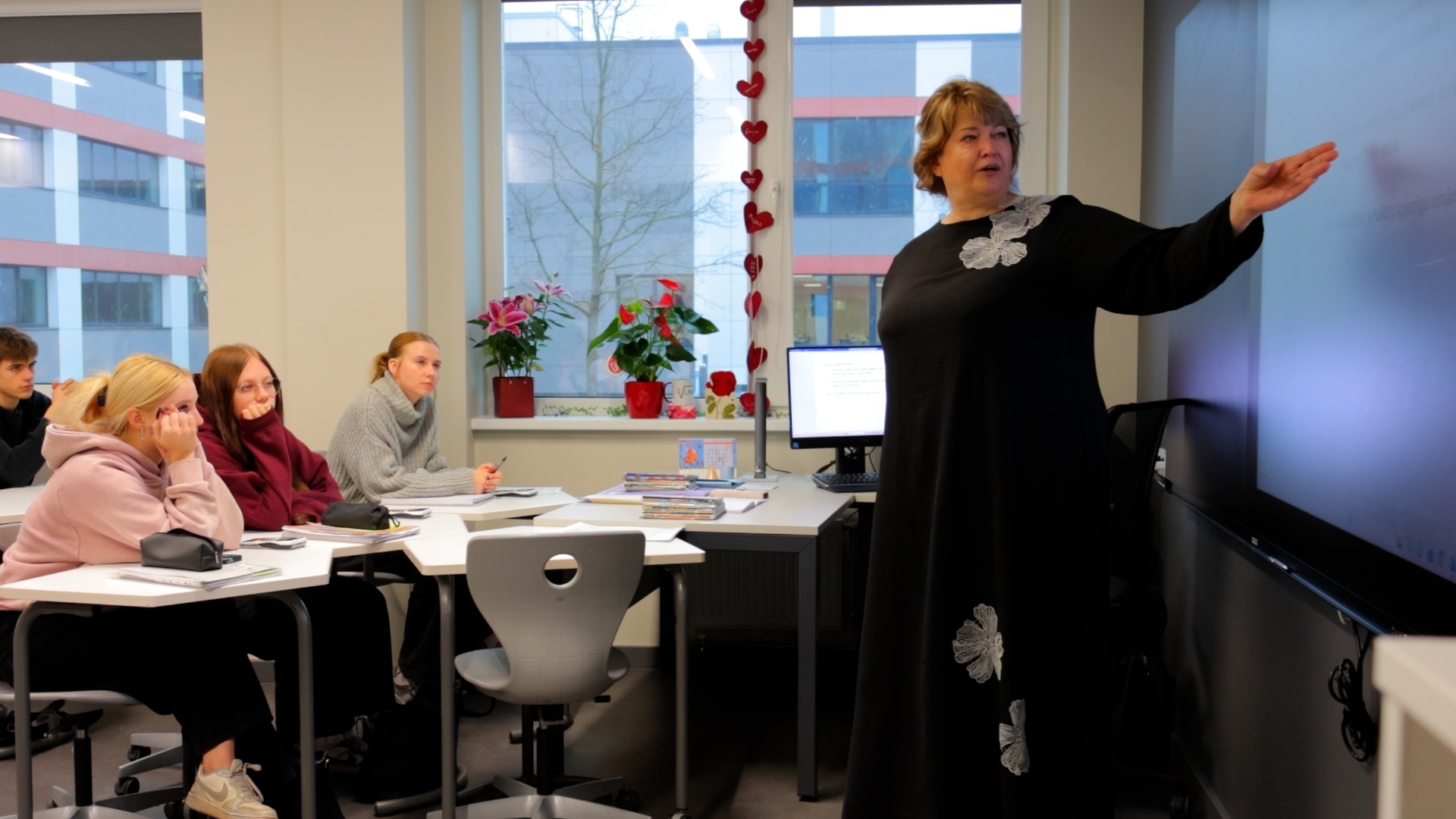 Skolotāja vada stundu modernā klasē, skolēni klausās un veic pierakstus.