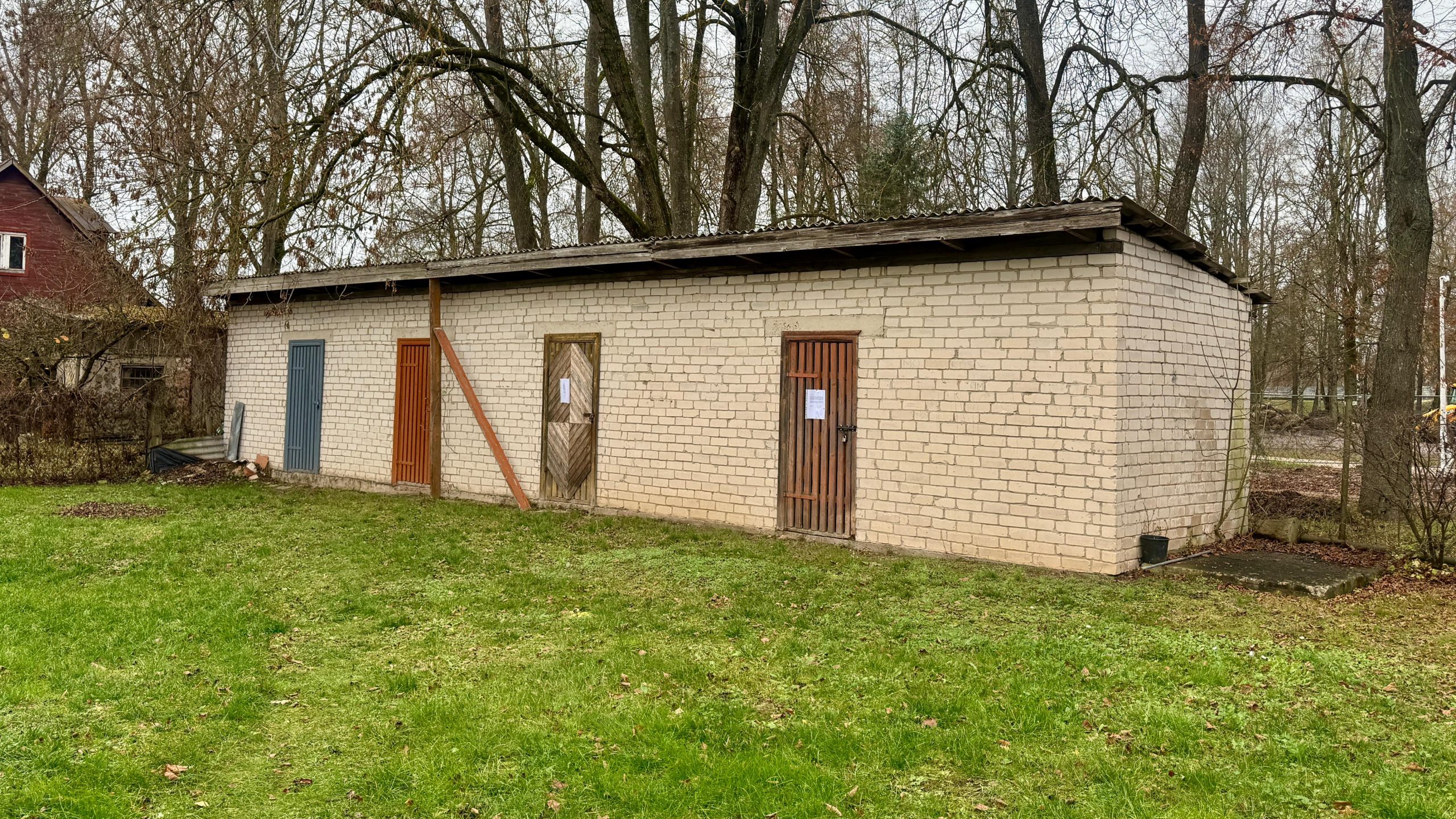Vispārējs skats uz zemu, gaišu ķieģeļu ēku ar vairākām atšķirīgām durvīm (šķūnīšiem), kas atrodas uz zaļa zāliena.