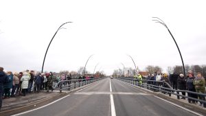 Panorāmas skats uz jauno, atklāto tiltu pār Salacas upi ar pulcētiem cilvēkiem gar abām margām.