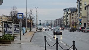 Jelgavas ielas skats ar automašīnām, autobusa pieturu un gājēju drūmā ziemas dienā.