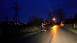 Gājējs tumsā iet pa piepilsētas ceļu ar ielu apgaismojumu un auto tālumā.