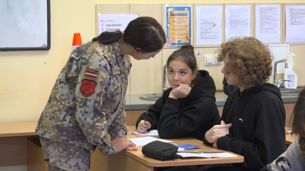 VAM instruktore militārā formā sarunājas ar diviem skolēniem pie galda.