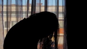 Sievietes siluets pie loga, noliektu galvu, attēlojot emocionālu spriedzi vai vardarbības sekas.