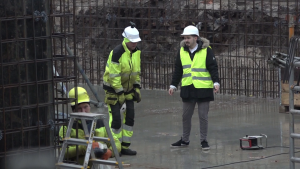 Būvniecības darbinieki ar aizsargķiverēm un atstarojošām vestēm strādā uz betona pamata pie armatūras konstrukcijas.