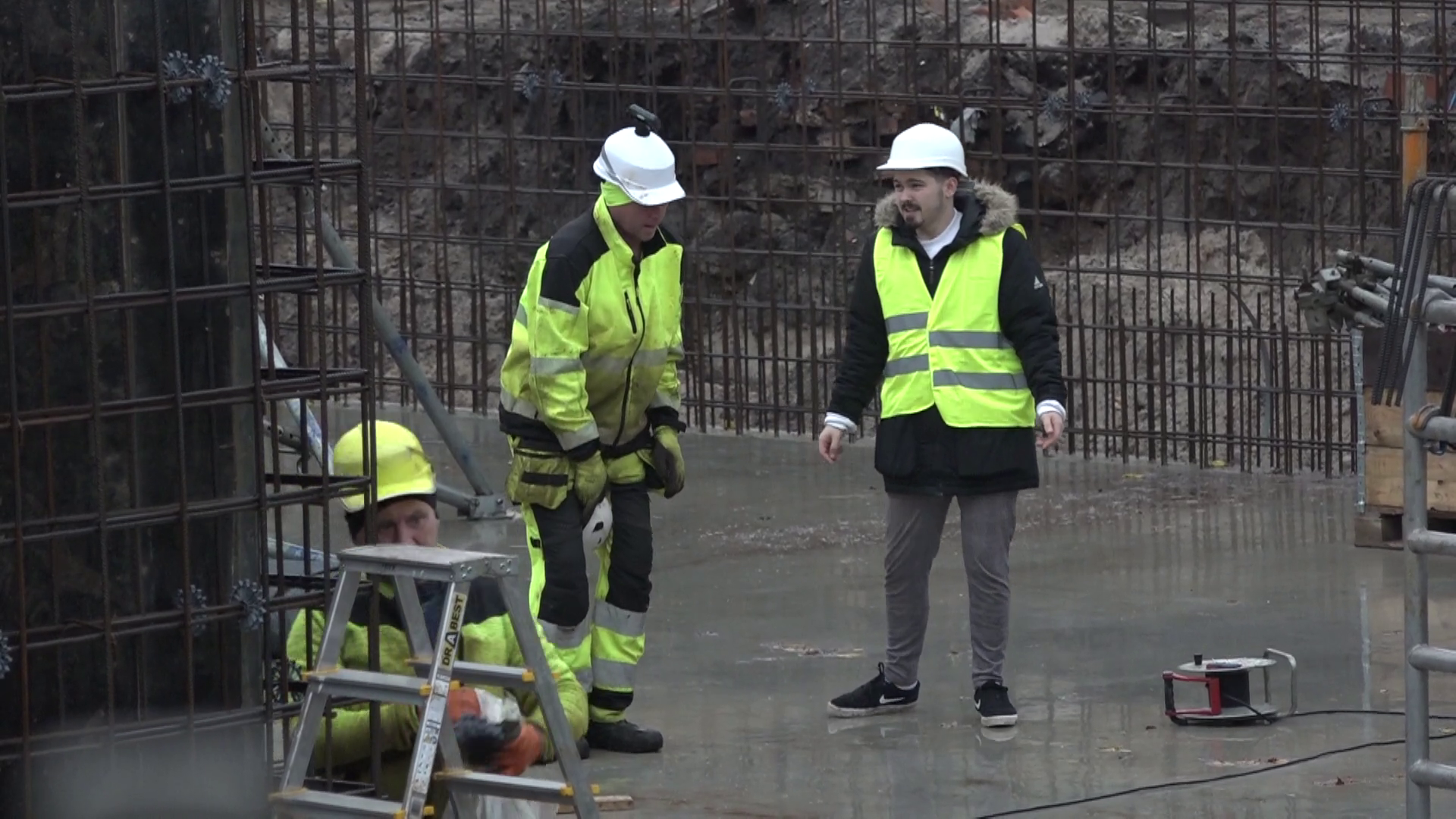 Būvniecības darbinieki ar aizsargķiverēm un atstarojošām vestēm strādā uz betona pamata pie armatūras konstrukcijas.