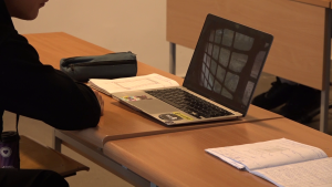 Students sēž pie galda ar atvērtu klēpjdatoru, burtnīcu un pildspalvu, strādājot mācību laikā.