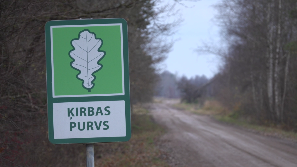 Ceļa zīme ar uzrakstu "Ķirbas Purvs" mežainā apvidū.