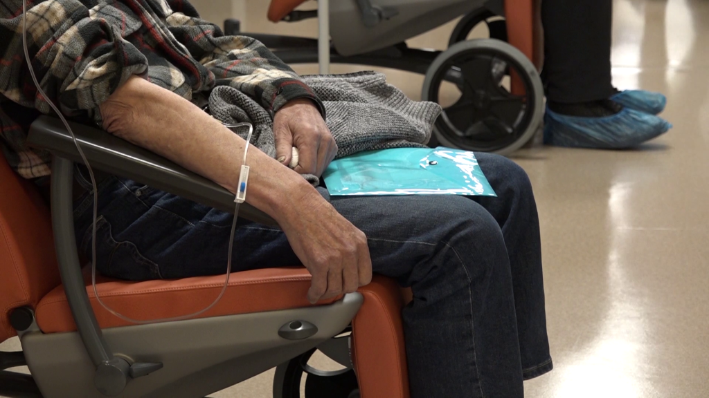 Tuvplāns uz cilvēka roku un kāju daļu, sēžot oranžā krēslā, ar intravenozu sistēmu, kas ievadīta rokā.