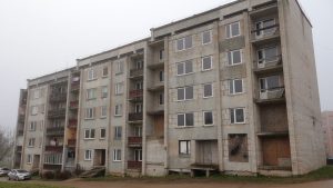 Pelēks, vecs piecstāvu daudzdzīvokļu nams ar vairākiem tukšiem logiem un aizbāztiem logiem/balkoniem ar koka plāksnēm.