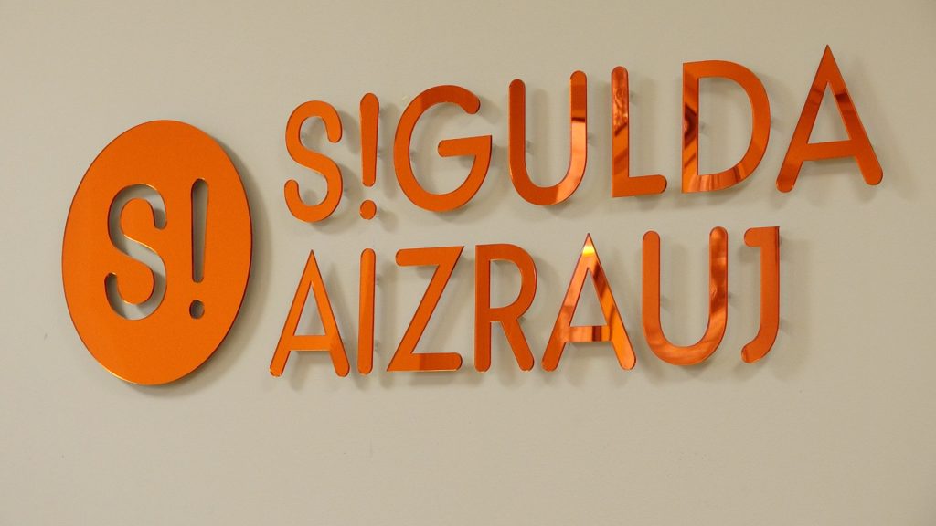 Tuvplāns uz liela, spilgti oranža uzraksta "S!GULDA AIZRAUJ" uz gaišas sienas.