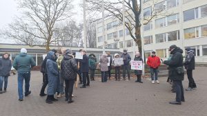 Apvidus skats uz aptuveni piecdesmit piketētājiem, kuri ar plakātiem pulcējas pie Jūrmalas domes ēkas ziemīgā dienā.