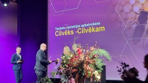 Apbalvošanas ceremonija uz skatuves ar violetu fona ekrānu. Ekrānā redzams uzraksts "Tieslietu ministrijas apbalvošana Cilvēks -Cilvēkam". Priekšplānā cilvēki un liels ziedu dekors.