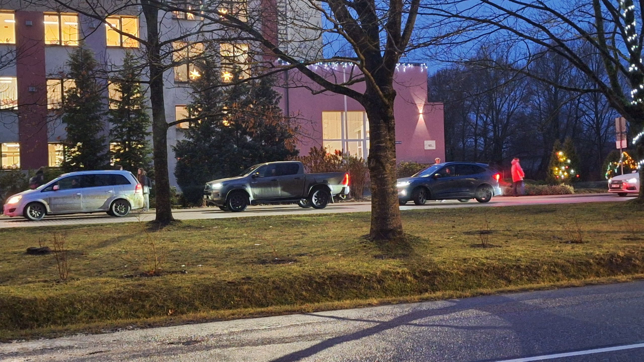 Agrā rīta stundā pie skolas ēkas, kas rotāta ar svētku gaismiņām, privātās automašīnas izlaiž skolēnus.