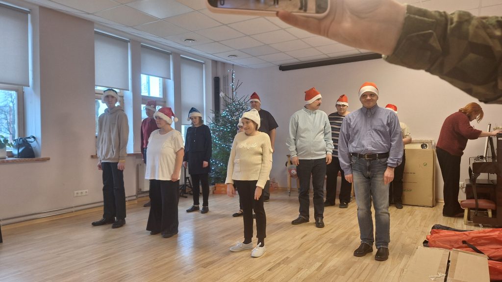 Aprūpes centra klienti svētku cepurēs gatavojas priekšnesumam telpā pie eglītes.