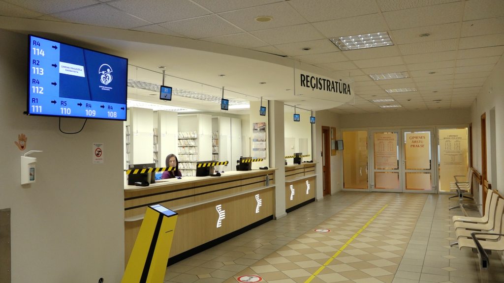 Plaša slimnīcas reģistratūra ar garu balta un dzeltena koka leti. Virs letes ir uzraksts "REĢISTRATŪRA" un rindas vadības ekrāns.
