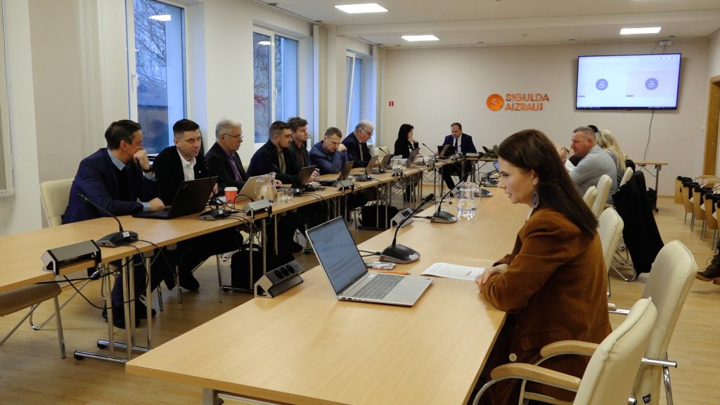Liela grupa deputātu sēž ap sanāksmju galdu, notiek domes sēde Siguldas novadā.