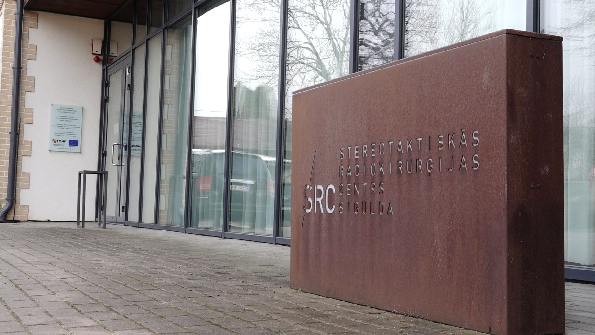 Rūsganas krāsas, liela taisnstūra izkārtne ar uzrakstu "STEREOTAKTISKĀS RADIOĶIRURĢIJAS CENTRS SRC SIGULDA" pie moderna stiklotas ēkas.