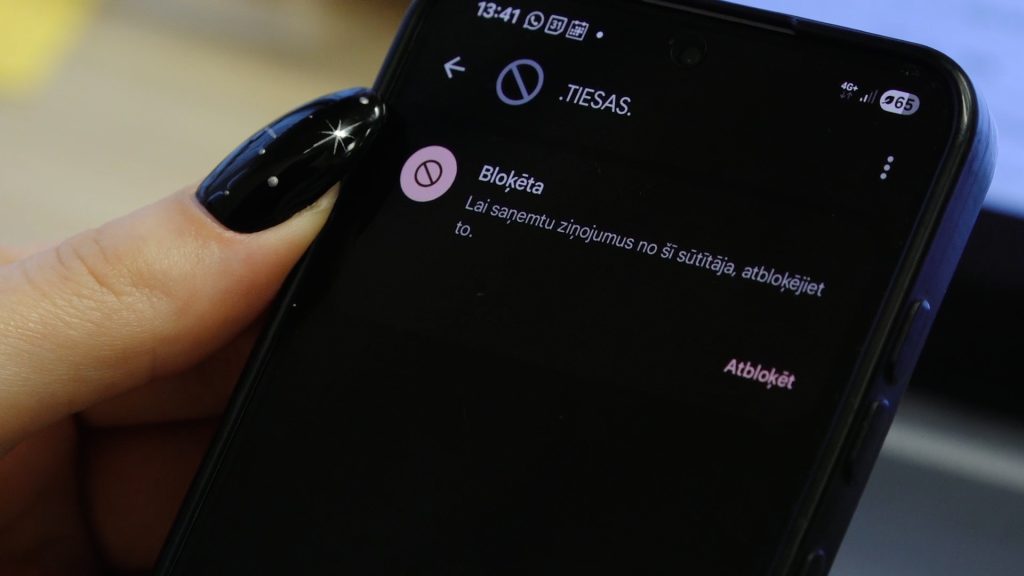Mobilā tālruņa ekrāns tuvplānā ar paziņojumu latviešu valodā par bloķētu sūtītāju.