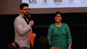 Pasākuma vadītājs ar mikrofonu uzrunā auditoriju, blakus stāv pasākuma dalībniece zaļā kleitā.