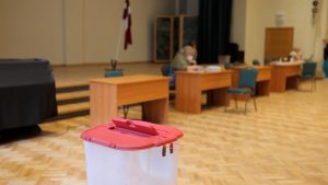 Balsošanas urna vēlēšanu iecirknī ar vēlēšanu komisijas locekļiem fonā