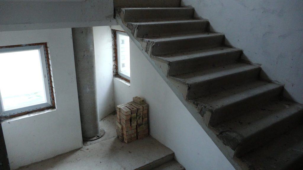 Puspabeigta betona kāpņu telpa ar tukšām sienām un logiem, un kaudzi ķieģeļu.