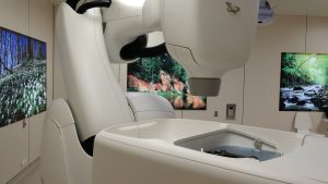 Tuvplāns uz baltu "CyberKnife" iekārtu ar melnu robotizētu roku un pacienta galdu. Fonā dabas attēli.