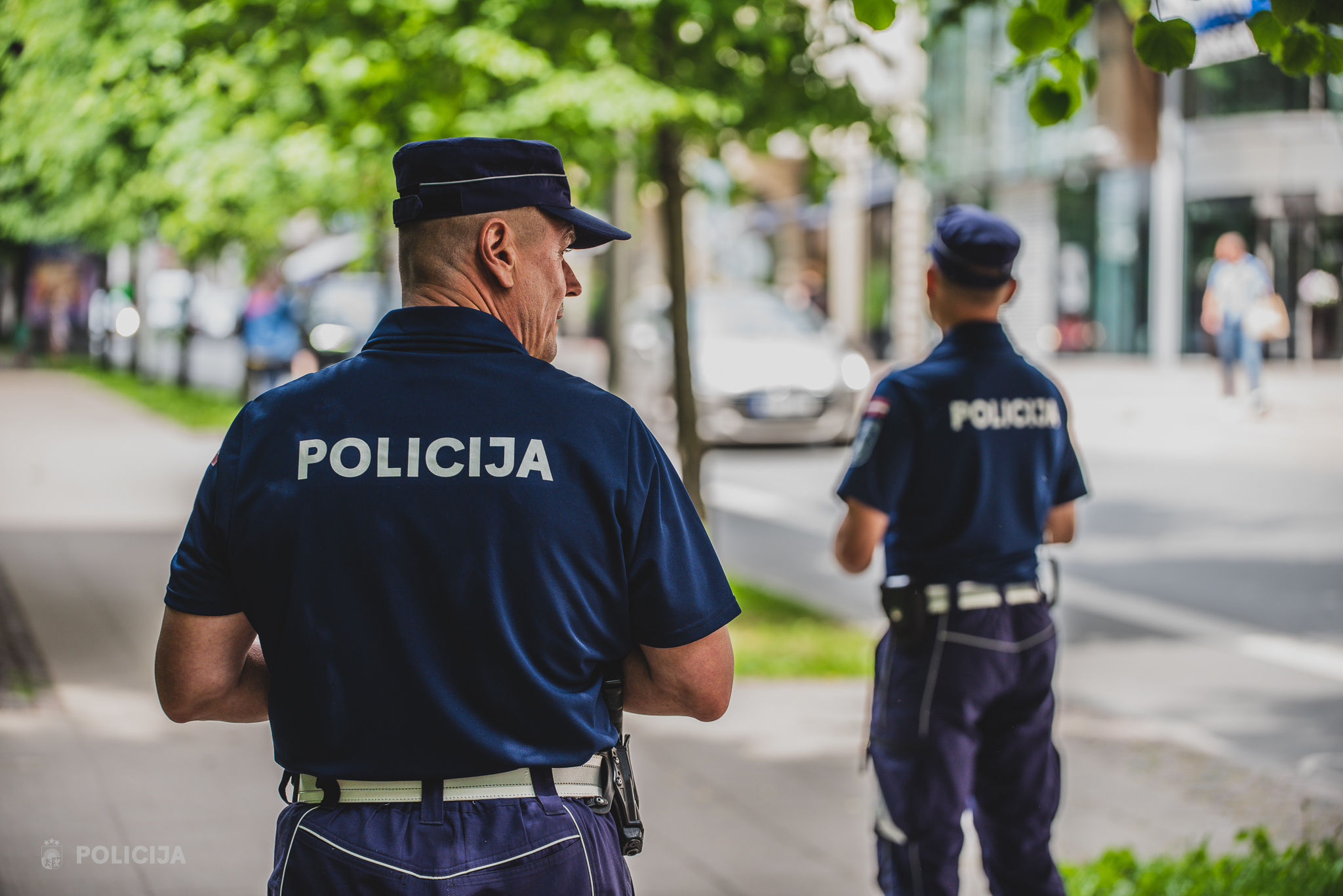 Divi policisti no mugurpuses patrulē pilsētas ielā vasaras dienā.