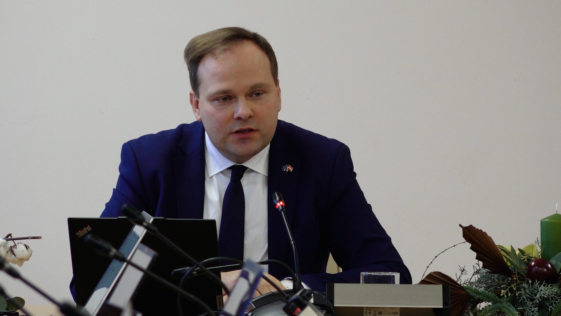 Linards Kumskis, tērpies tumšā uzvalkā un kaklasaitē, runā pie mikrofoniem Siguldas novada domes sēdē.
