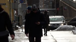 Gājēji ziemā pārvietojas pa pilsētas ietvi, viens skatās telefonā, fonā automašīnas