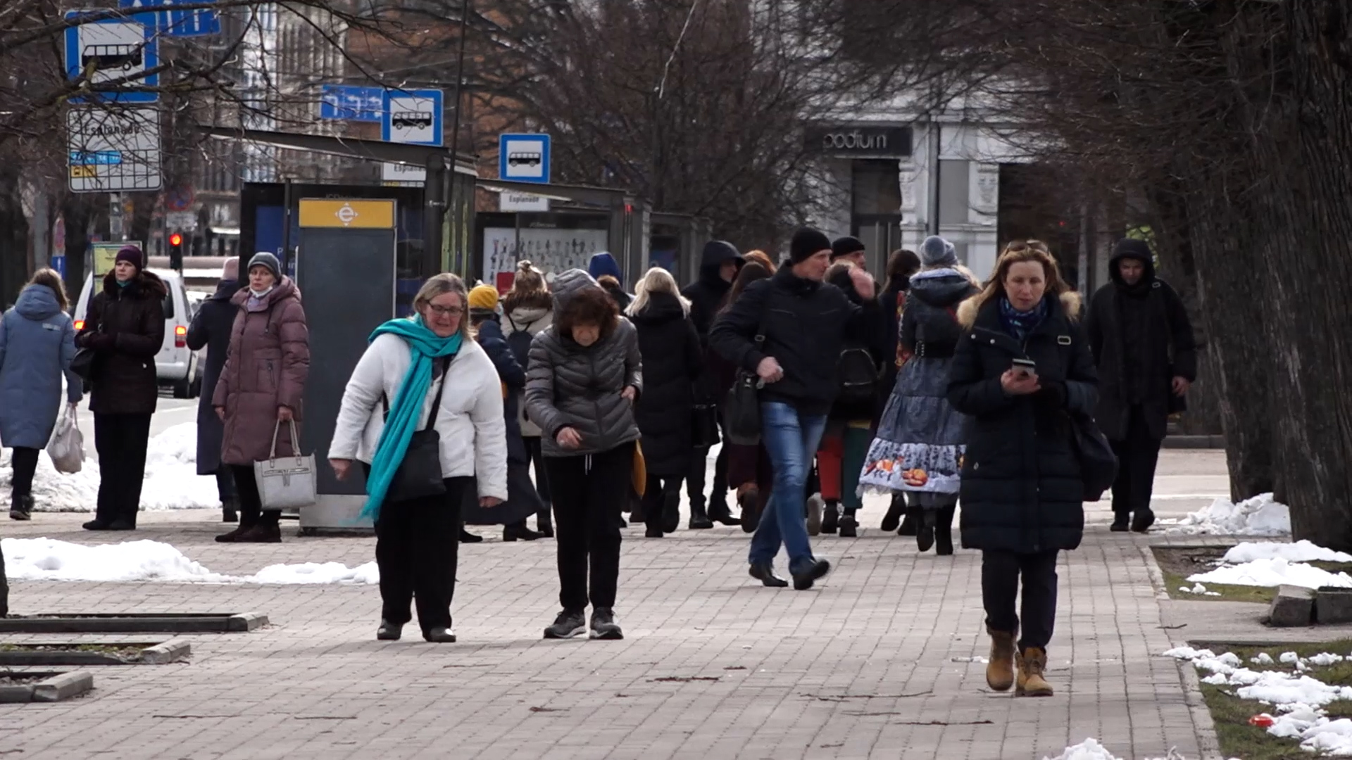 Cilvēki ziemā iet pa pilsētas ietvi pie sabiedriskā transporta pieturas
