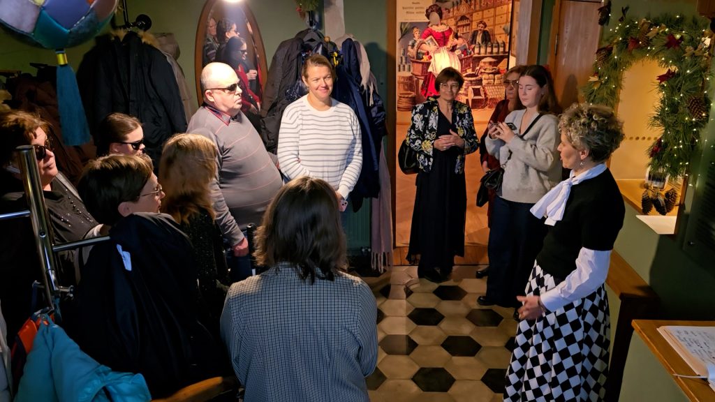 Apmeklētāju grupa un gide pulcējas muzeja telpā, kurā redzami svētku rotājumi un mēteļu pakaramie.