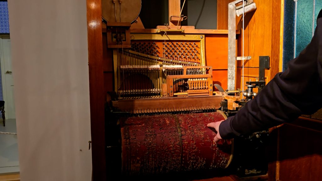 Tuvplāns ar 20. gadsimta sākuma orkestriona mehānismu un cilvēka roku, kas pieskaras perforētajam rullim.