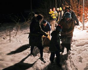 Cilvēki, tostarp bērni, sniegā velk bluķi ziemas saulgriežu tradīcijas laikā.